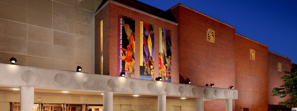 Exterior view of the Jorgensen Center at dusk, featuring a brick and concrete facade with colorful vertical banners and illuminated entrance doors where people are entering.
