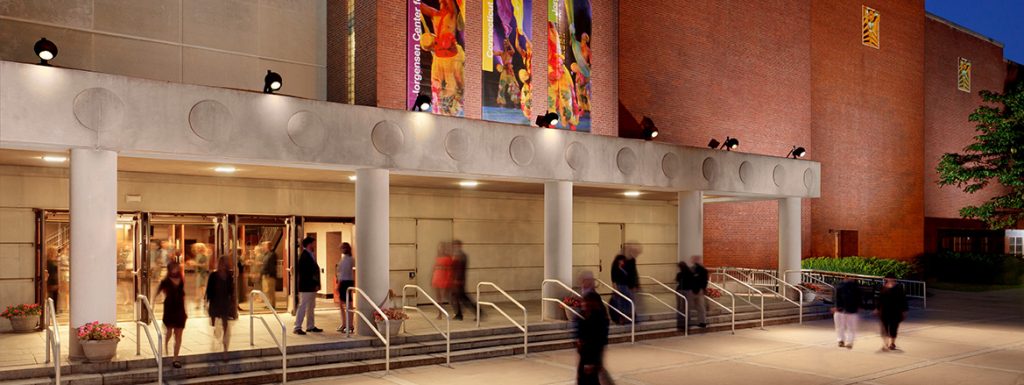 Exterior view of the Jorgensen Center at dusk, featuring a brick and concrete facade with colorful vertical banners and illuminated entrance doors where people are entering.