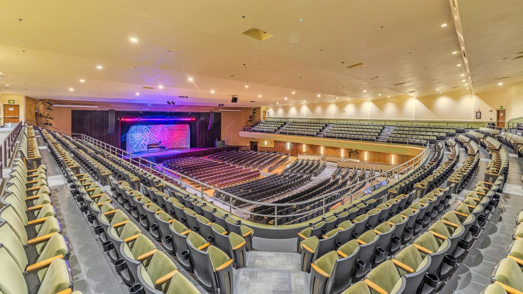 Wide view of the Jorgensen Center for the Performing Arts auditorium, showing rows of empty seats on the main floor and balcony facing a lit stage with colorful blue and red lighting.