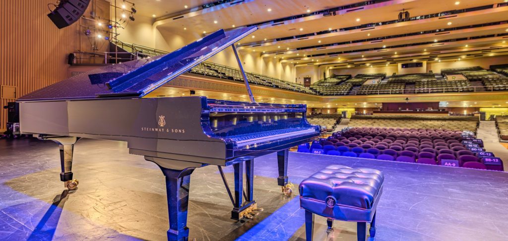 A Steinway & Sons grand piano sits on stage under warm lighting in an empty concert hall, with rows of seats visible in the background.