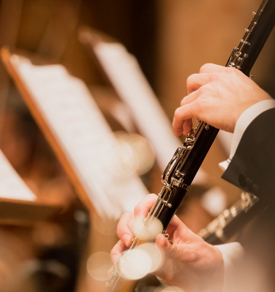 A musician in formal concert attire plays a clarinet during an orchestral performance, with sheet music blurred in the background.
