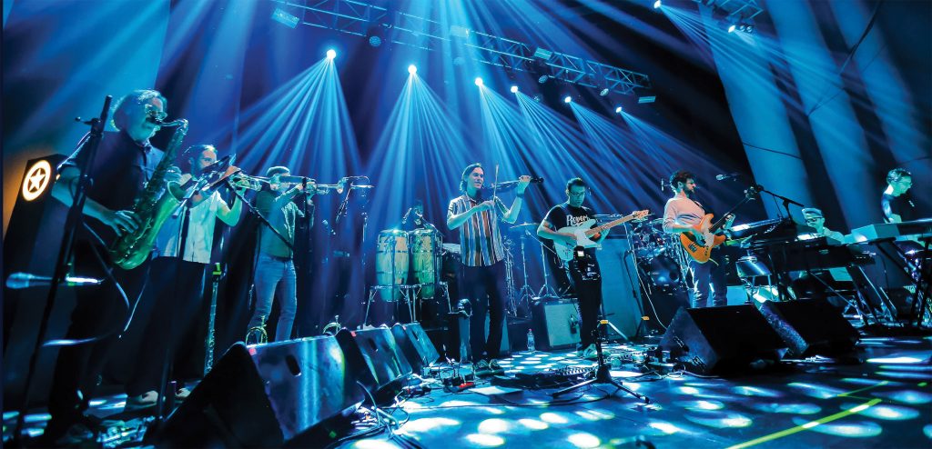 A band performs live on stage under bright blue and purple lights, featuring multiple musicians playing saxophone, trombone, violin, electric guitar, bass, keyboards, and percussion instruments.