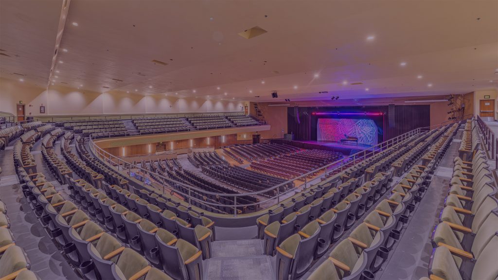 Wide view of the Jorgensen Center for the Performing Arts auditorium, showing rows of empty seats on the main floor and balcony facing a lit stage with colorful blue and red lighting.