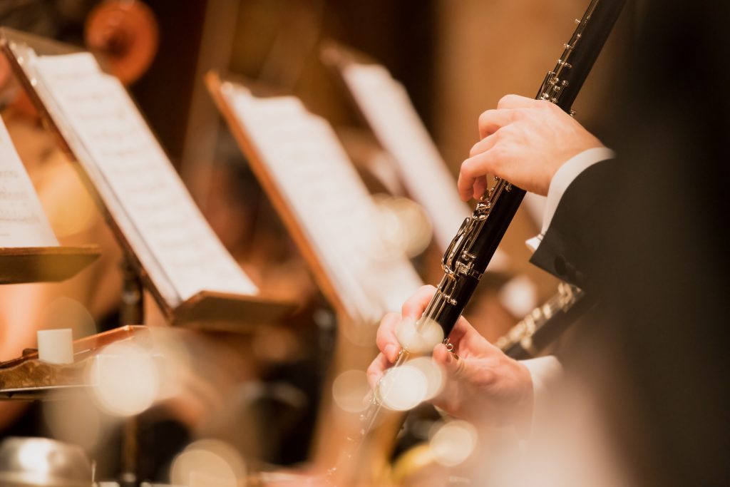 A musician in formal concert attire plays a clarinet during an orchestral performance, with sheet music blurred in the background.
