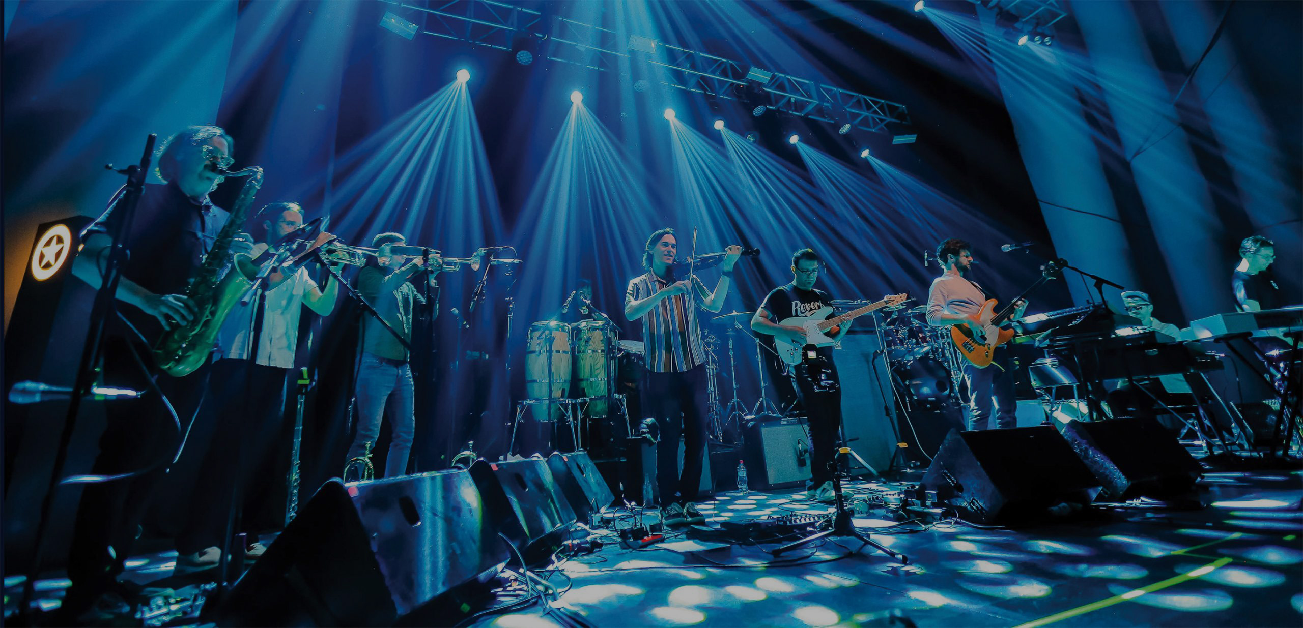 A band performs live on stage under bright blue and purple lights, featuring multiple musicians playing saxophone, trombone, violin, electric guitar, bass, keyboards, and percussion instruments.